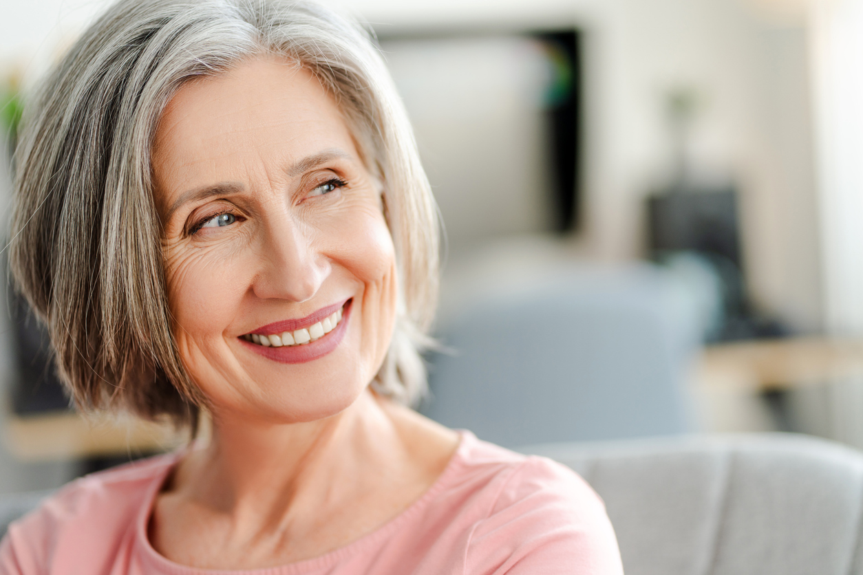 From Gaps to Great Smiles: How Dental Implants Transform Confidence, Closeup portrait smiling confident senior woman sitting on comfortable sofa at home. Happy retired female with stylish gray hair, white teeth looking away. Natural beauty, healthy lifestyle concept