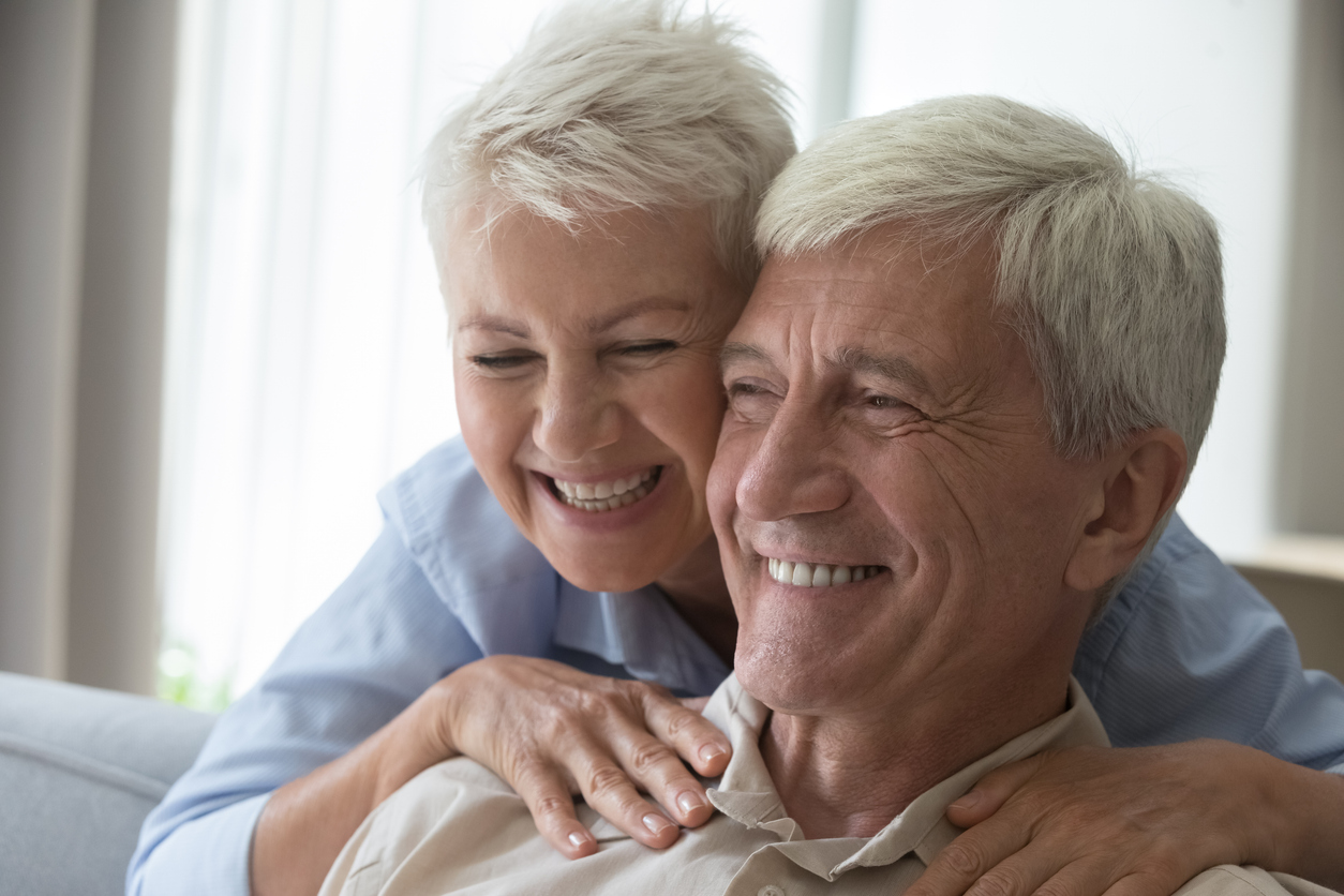 Dental Implants vs. Removable Implant Dentures: Which Is the Better Long-Term Solution?, Close up shot affectionate smiling middle aged wife embrace husband pensioner from behind massage shoulders of beloved man express love care devotion. Romantic cheerful old age couple hug touch heads